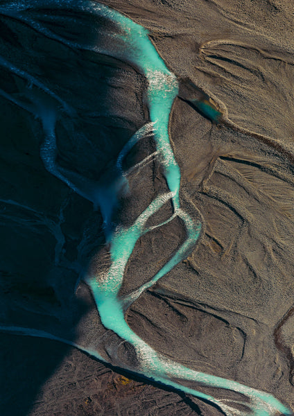 Aerial view of a winding turquoise river cutting through dark brown and black sedimentary terrain, creating branching channels and patterns of light and shadow.